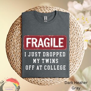May include: Dark heather gray t-shirt with the word "FRAGILE" in a red rectangle, and the text "I JUST DROPPED MY TWINS OFF AT COLLEGE" in white. The shirt is displayed on a woven surface.