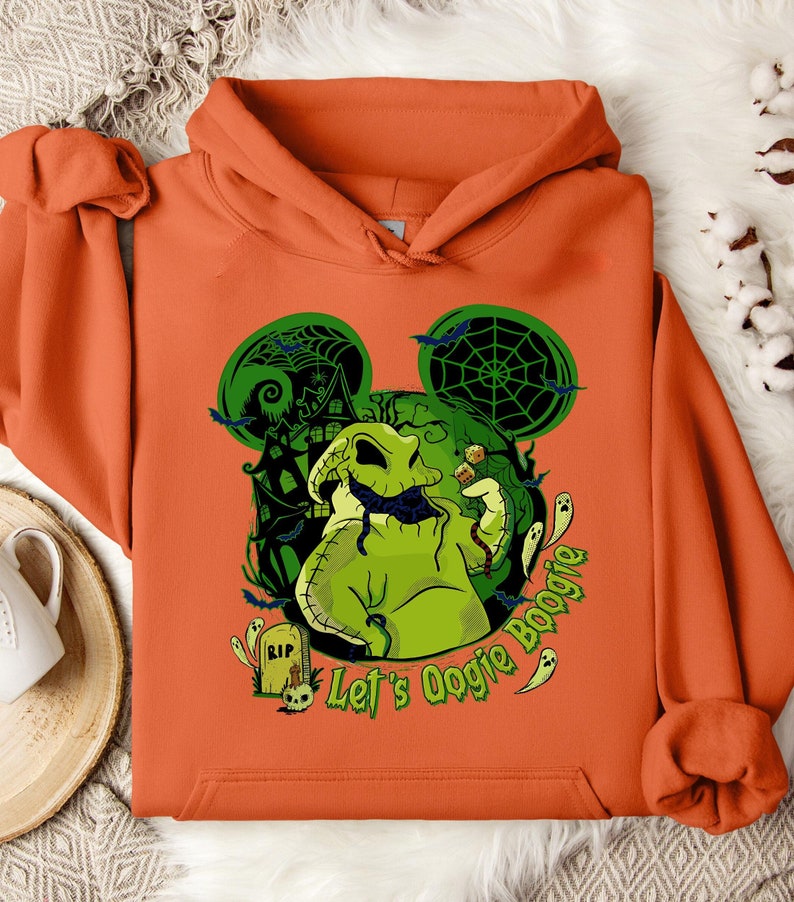 May include: Orange hoodie featuring a green Oogie Boogie design with a haunted house, bats, and the text "Let's Oogie Boogie." The design includes a tombstone and dice. The hoodie is made of a soft material.