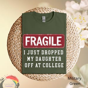 May include: Military green t-shirt with the word "FRAGILE" in a red rectangle. Below the rectangle, the text reads "I JUST DROPPED MY DAUGHTER OFF AT COLLEGE". The shirt is displayed on a woven surface.