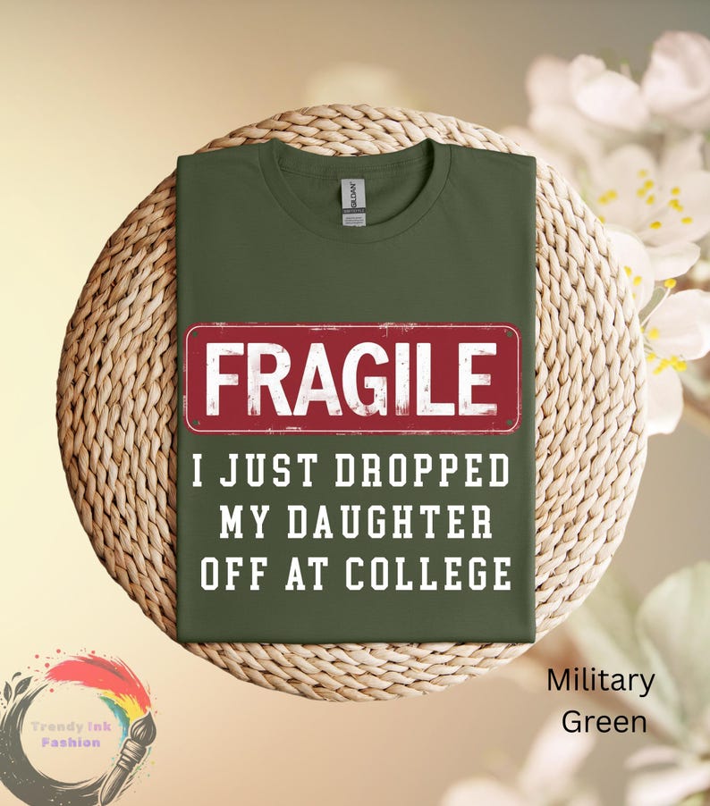 May include: Military green t-shirt with the word "FRAGILE" in a red rectangle. Below the rectangle, the text reads "I JUST DROPPED MY DAUGHTER OFF AT COLLEGE". The shirt is displayed on a woven surface.