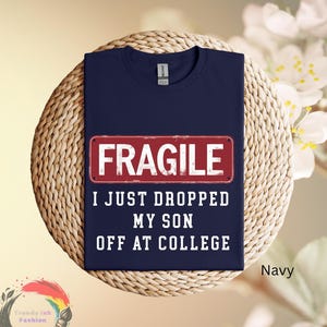 May include: Navy blue t-shirt with the word "FRAGILE" in white block letters on a red rectangle. Below, the text reads "I JUST DROPPED MY SON OFF AT COLLEGE". The shirt is displayed on a woven surface.