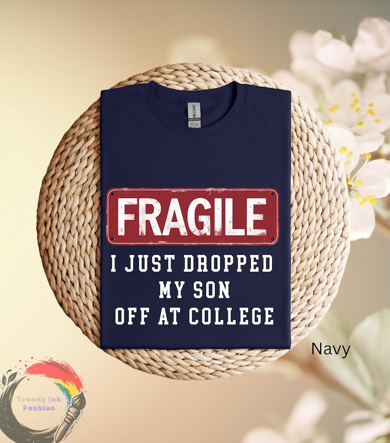 May include: Navy blue t-shirt with the word "FRAGILE" in white block letters on a red rectangle. Below, the text reads "I JUST DROPPED MY SON OFF AT COLLEGE". The shirt is displayed on a woven surface.