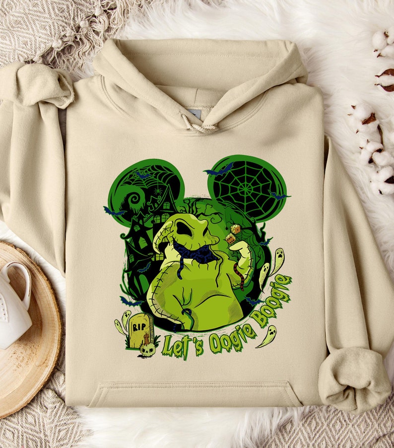 May include: Beige hoodie featuring a green Oogie Boogie design. The design includes a green Oogie Boogie character, bats, a haunted house, and the text "Let's Oogie Boogie". The hoodie has a drawstring hood and a kangaroo pocket.