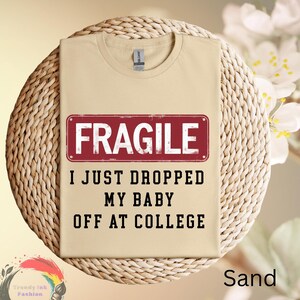 May include: Sand-colored t-shirt with the text "FRAGILE" in a red rectangle, and the phrase "I JUST DROPPED MY BABY OFF AT COLLEGE" in black. The shirt is displayed on a woven surface.