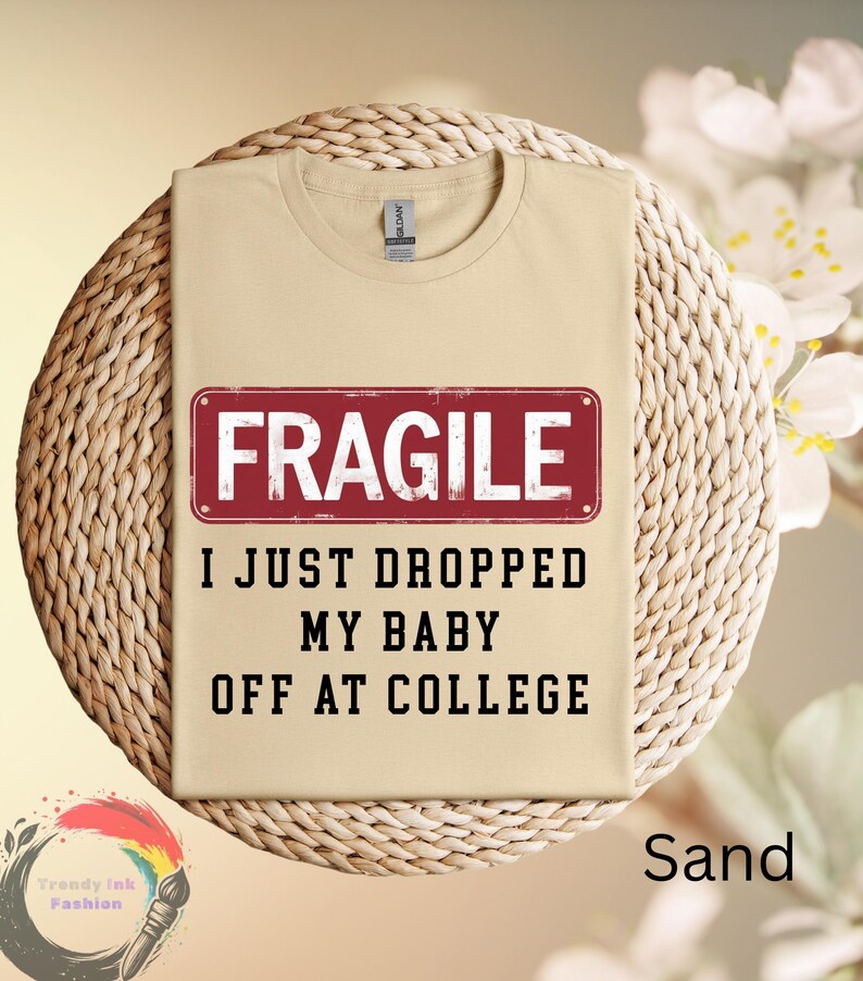 May include: Sand-colored t-shirt with the text "FRAGILE" in a red rectangle, and the phrase "I JUST DROPPED MY BABY OFF AT COLLEGE" in black. The shirt is displayed on a woven surface.