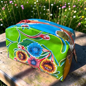 May include: A rectangular zippered pouch featuring a vibrant green base with a colorful floral pattern in shades of blue, yellow, and red. The top panel is bright blue, and the zipper is a contrasting red. The pouch has a smooth, coated finish.