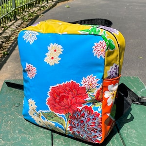 May include: A blue, yellow and orange backpack with a floral print. The backpack has a zip closure and a black strap.