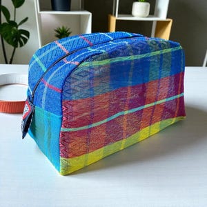 May include: A rectangular, zippered cosmetic bag with a vibrant plaid pattern. The bag features a color palette of blue, red, yellow, and green. The bag is made of a woven material and has a black zipper.