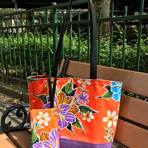 K&ouml;nnte beinhalten: Orangefarbene Einkaufstasche mit Blumendruck und lilafarbenem Boden. Die Tasche hat einen schwarzen Henkel und eine kleine passende Tasche.