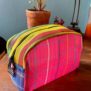 以下が含まれることがあります： 黄色と緑のストライプのジッパーが付いたピンクとブルーの格子柄のメイクアップバッグ。バッグには「The Shine」と書かれた小さなタグが付いています。