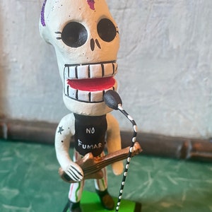 May include: A white skull figurine with a purple mohawk, wearing a black shirt with the text "NO FUMAR" and holding a guitar. The figurine is standing on a green base with a black microphone stand.