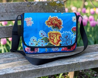 Könnte beinhalten: Eine leuchtend blaue Umhängetasche mit einem lebendigen Blumenmuster aus großen gelben, orangefarbenen und roten Pfingstrosen sowie kleineren weißen und blauen Blüten. Die Tasche hat einen schwarzen Klappenrand, schwarze Seitenteile und einen breiten schwarzen Riemen.