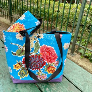 K&ouml;nnte beinhalten: Eine blaue und wei&szlig;e Blumentasche mit schwarzen Tr&auml;gern. Die Tasche hat ein lila Futter und ein Blumenmuster.
