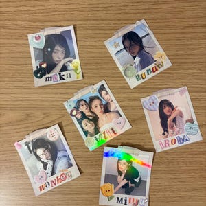Peut inclure: Six tirages photo carrés présentant des portraits de personnes, chacun décoré de petits autocollants colorés et de ruban adhésif. Les tirages incluent les noms Moka, Yunah, Iroha, Wonhee et Minju en lettres colorées, avec un tirage montrant un groupe.
