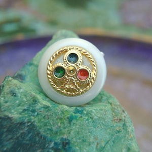 May include: A white button with a gold design and four colored gemstones. The gemstones are green, red, and gold.