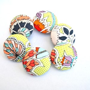 Puede incluir: Un conjunto de seis botones de tela coloridos con una variedad de patrones florales y geométricos. Los botones son redondos y tienen un fondo amarillo con un contorno blanco.