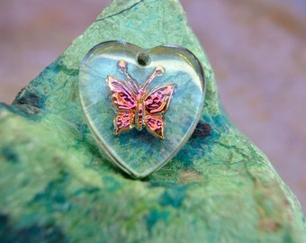 Puede incluir: Un colgante transparente en forma de corazón con un diseño de mariposa rosa y dorada en su interior. El colgante está sobre una roca verde y marrón.