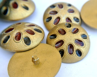 May include: A close-up of five gold-colored buttons with a raised, textured surface. The buttons have a circular design with a ring of red and black oval-shaped accents.