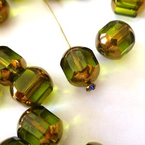 May include: Close-up of faceted olive green and bronze-colored beads scattered on a white surface. Some beads are strung on a gold-colored wire. A small blue bead is at the end of the wire. These beads are suitable for jewelry making.