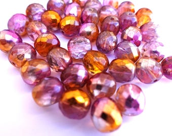 May include: A collection of faceted beads in shades of purple, pink, and gold. The beads have a shiny, iridescent finish and are scattered across a white surface. These beads are suitable for jewelry making and crafting projects.