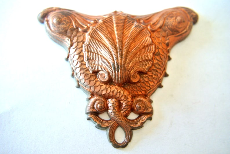 May include: A copper-colored metal ornament shaped like two fish flanking a seashell. The ornament has a detailed design with scales and a textured surface.
