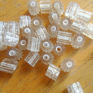 May include: A collection of clear, rectangular beads with a striped, glittery design. Each bead has a white, circular hole in the center. The beads are scattered on a wooden surface, showcasing their translucent quality.
