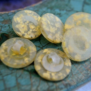 May include: Five round, translucent buttons with a gold fleck design. The buttons are arranged on a blue and green textured surface.