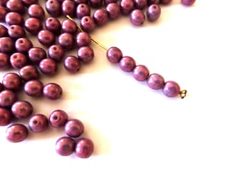 May include: Plum-coloured beads with a metallic sheen, some strung on gold wire, others scattered on a white surface. These round beads are suitable for jewellery making. The beads are approximately 0.8 cm in diameter.