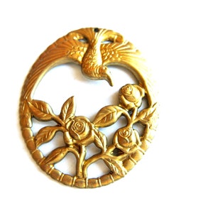 Könnte beinhalten: Ein goldfarbener Metallschild mit einem stilisierten Vogel mit ausgebreiteten Flügeln und einem Blumendesign mit Rosen. Das Schild ist rund und hat ein filigranes, kunstvolles Design.
