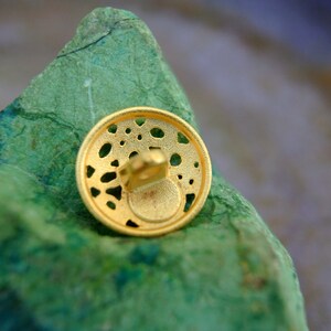 K&ouml;nnte beinhalten: Ein goldfarbener Metallknopf mit einem dekorativen, filigranen Design. Der Knopf hat ein kleines Loch in der Mitte zum N&auml;hen.