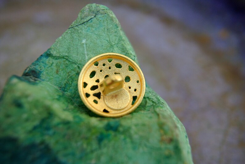 K&ouml;nnte beinhalten: Ein goldfarbener Metallknopf mit einem dekorativen, filigranen Design. Der Knopf hat ein kleines Loch in der Mitte zum N&auml;hen.