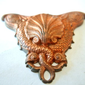 May include: A copper-colored metal ornament shaped like two fish flanking a seashell. The ornament is detailed with scales and swirls.