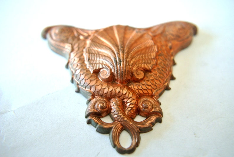 May include: A copper-colored metal ornament shaped like two fish flanking a seashell. The ornament is detailed with scales and swirls.
