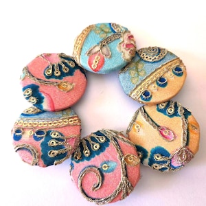 May include: A collection of seven round, decorative buttons. The buttons feature a pink, blue, and gold color scheme with intricate embroidered designs. The buttons are arranged in a circular pattern against a white background.
