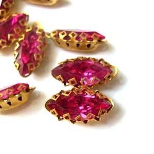 May include: A group of pink glass rhinestones with gold metal settings. The rhinestones are in the shape of a marquise and have a faceted design.