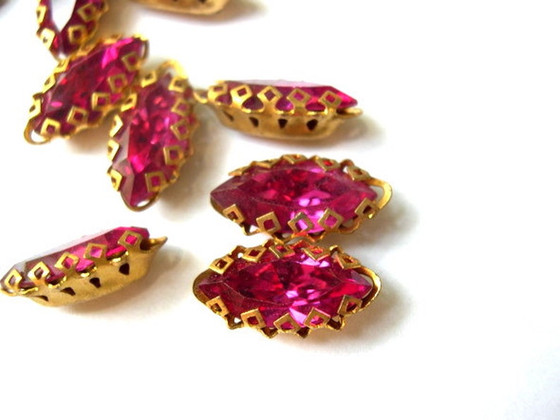 Puede incluir: Un grupo de piedras de cristal rosa con engastes de metal dorado. Las piedras de cristal tienen forma de marquesa y un dise&ntilde;o facetado.