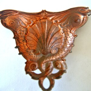 May include: A copper-colored metal ornament shaped like two seahorses with their tails intertwined, forming a shell-like shape in the center.
