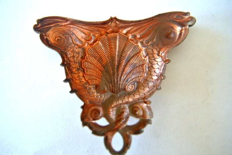 May include: A copper-colored metal ornament shaped like two seahorses with their tails intertwined, forming a shell-like shape in the center.