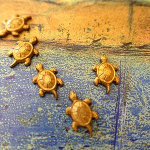 May include: Five small, gold-coloured turtle-shaped charms or buttons. Each turtle features a detailed, raised shell pattern with a central hexagonal plate surrounded by smaller segments, four legs, and a tail, arranged in a scattered formation.