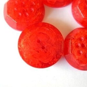 Pu&ograve; includere: Una collezione di bottoni rotondi rosso brillante con una consistenza traslucida e marmorizzata. Ogni bottone presenta una superficie liscia e lucida con due fori centrali per il fissaggio, mostrando un aspetto vivace e semitrasparente.