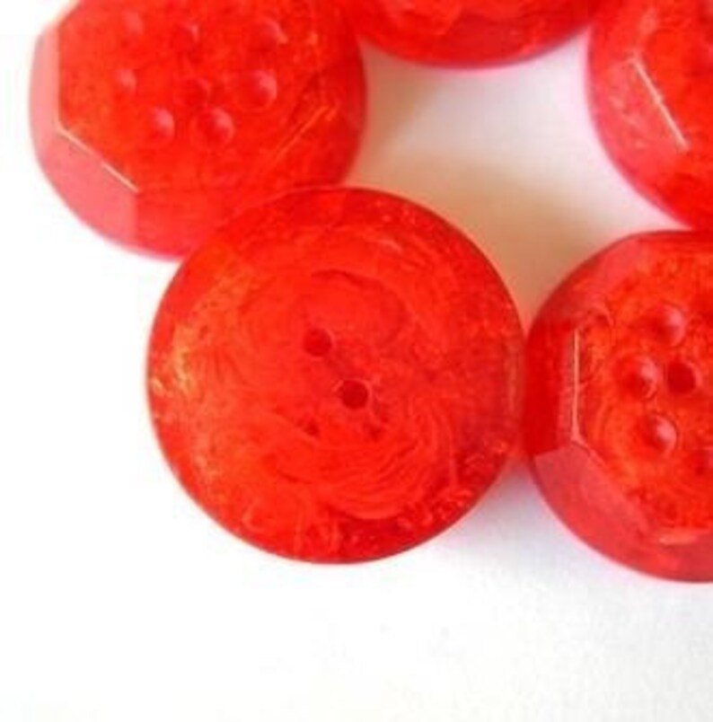 Pu&ograve; includere: Una collezione di bottoni rotondi rosso brillante con una consistenza traslucida e marmorizzata. Ogni bottone presenta una superficie liscia e lucida con due fori centrali per il fissaggio, mostrando un aspetto vivace e semitrasparente.