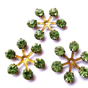 May include: Three gold-toned metal snowflake-shaped embellishments with green rhinestones. Each snowflake has six points with a rhinestone at each point.