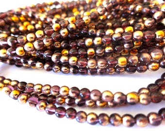 May include: Close-up of multiple strands of small, round beads in shades of purple, gold, and brown. The beads are strung together, creating a textured effect. These beads are likely used for jewellery making or other crafts.