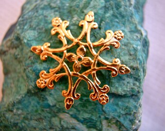 May include: A gold-colored metal snowflake ornament with intricate details. The snowflake is on a green and blue textured surface.