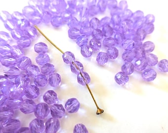 May include: A scattering of light purple, faceted beads on a white surface. A gold-coloured head pin has several beads threaded onto it. The small beads have a sparkling, crystal-like appearance, suitable for jewellery making.
