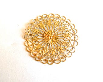 May include: A gold-colored, circular filigree ornament with a floral design. The intricate piece features a central starburst pattern surrounded by delicate leaf-like shapes and a scalloped edge. The ornament is approximately 4 inches in diameter.