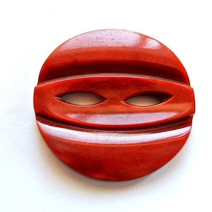 May include: A red plastic button with a stylized eye design. The button is round and has a glossy finish.