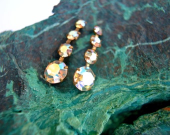 May include: A pair of gold earrings with five clear iridescent rhinestones on each earring. The earrings are on a green and brown textured surface.