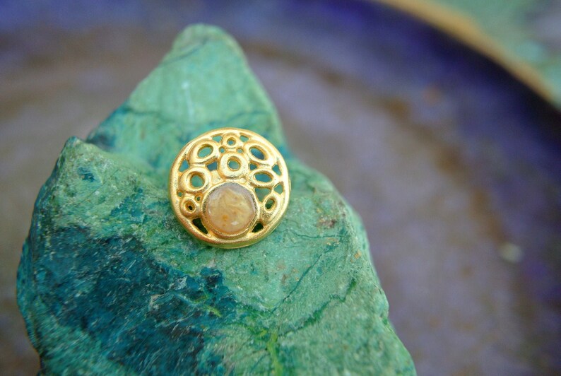 K&ouml;nnte beinhalten: Ein goldfarbener Ring mit einem runden Design, das einen kleinen, klaren Stein in der Mitte tr&auml;gt. Der Ring liegt auf einem gr&uuml;n-blauen Stein.
