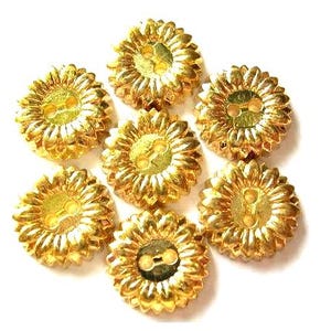 May include: A collection of seven circular gold-colored buttons arranged in a cluster. Each button features a textured, radiating petal design resembling a flower, with two small holes in the center for attachment to fabric or other materials.